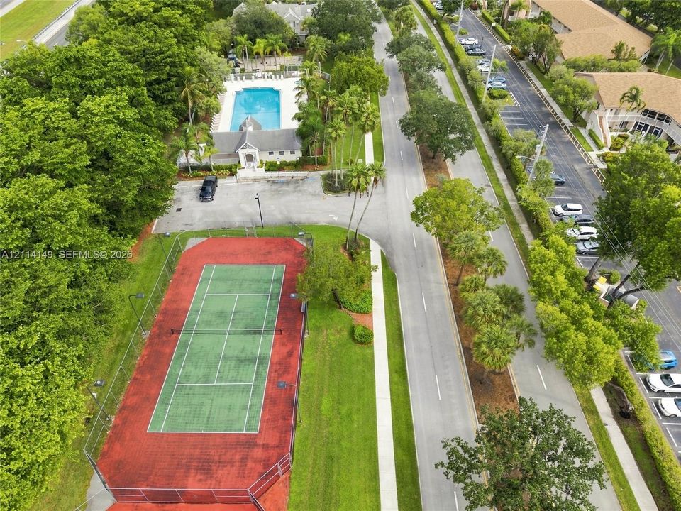 Aerial perspective featuring a fenced tennis court, paved parking areas, and a community swimming pool surrounded by mature palm trees and green space