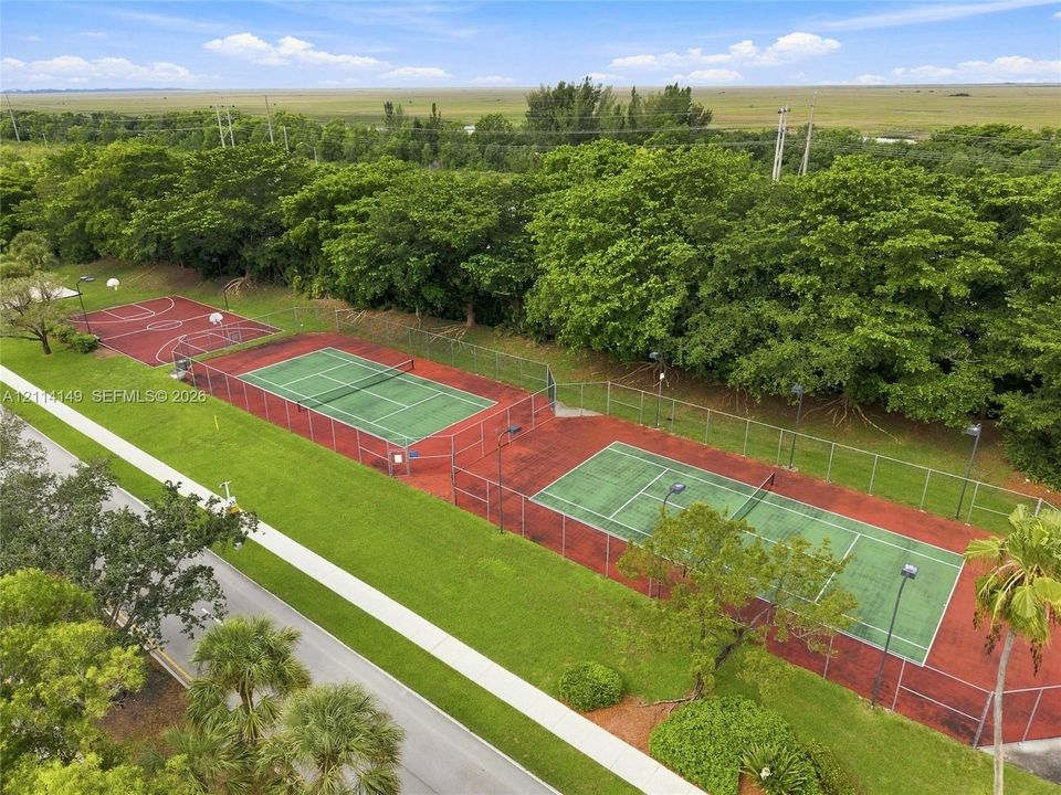 Outdoor recreation area featuring two fenced tennis courts, a basketball court, and mature landscaping