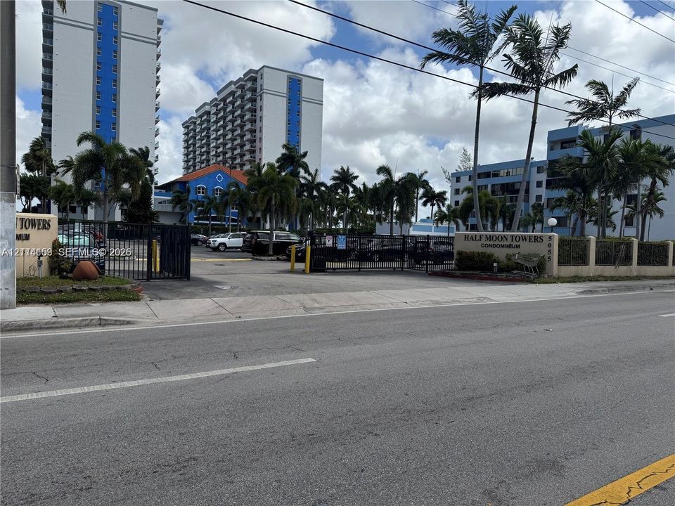 Half Moon Towers gated entrance featuring a security gate, stone masonry signage, and mature palm landscaping
