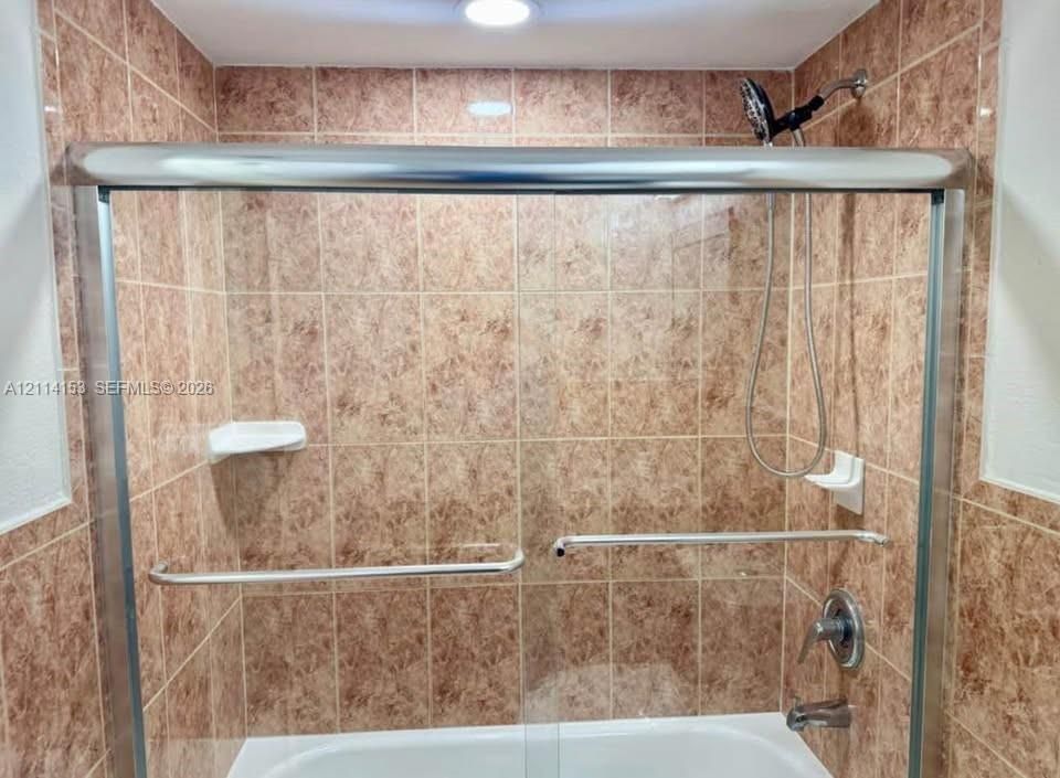 Bathtub and shower combination featuring floor-to-ceiling tile surround, sliding glass door enclosure, built-in corner shelving, and metallic hardware