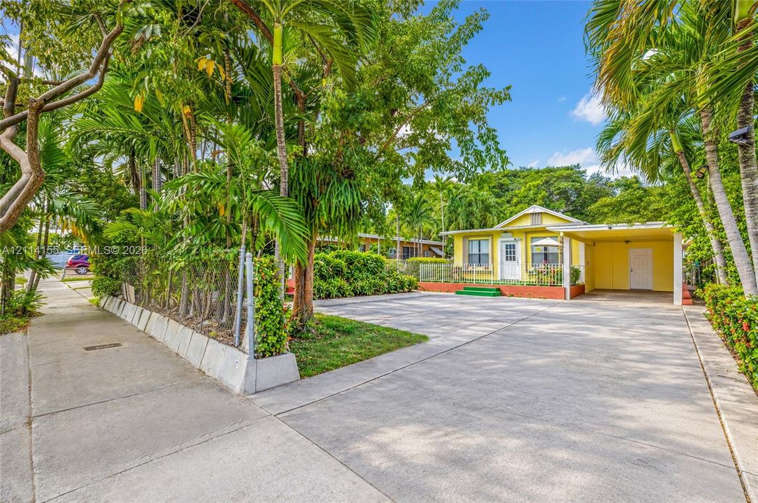Single-story residence featuring a paved driveway, attached carport, perimeter landscaping with mature palms, and a low-profile masonry retaining wall