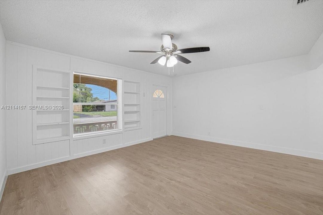 Interior space featuring wood-finish flooring, built-in shelving flanking a large window, a ceiling fan with integrated lighting, and a single-light panel entry door
