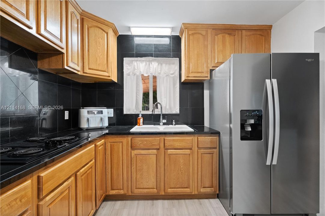 Kitchen featuring shaker-style wood cabinetry, black tile backsplash, integrated stainless steel gas cooktop, double-basin sink with a gooseneck faucet, and a side-by-side stainless steel refrigerator