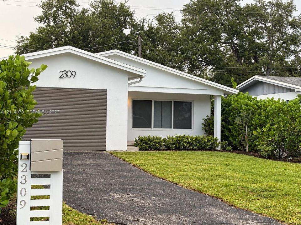 Single-story exterior featuring a white stucco facade, an attached one-car garage with a horizontal-slat door, a paved driveway, and a custom modern mailbox