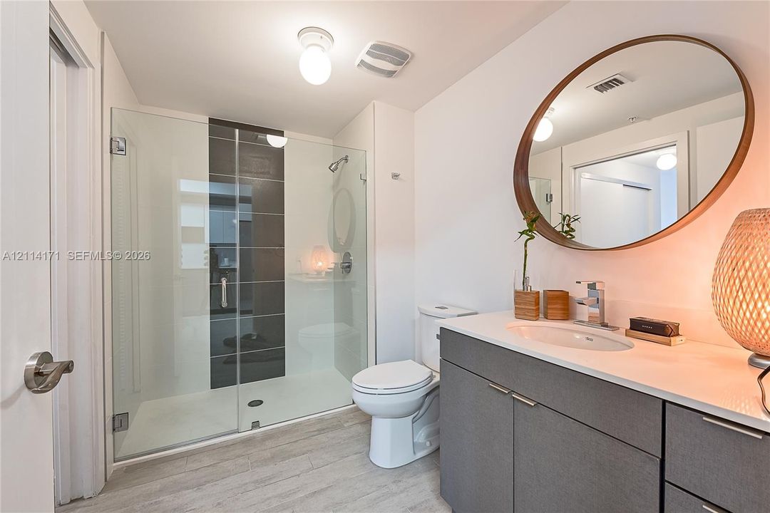 Bathroom featuring a glass-enclosed shower with a central dark tile accent, grey flat-panel vanity cabinetry, white countertop with an integrated oval sink, and a large oval wall-mounted mirror