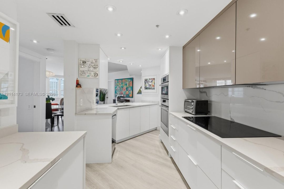 Modern kitchen featuring high-gloss white and metallic cabinetry, stone-finish countertops, full-height stone-finish backsplash, built-in wall oven, and recessed lighting