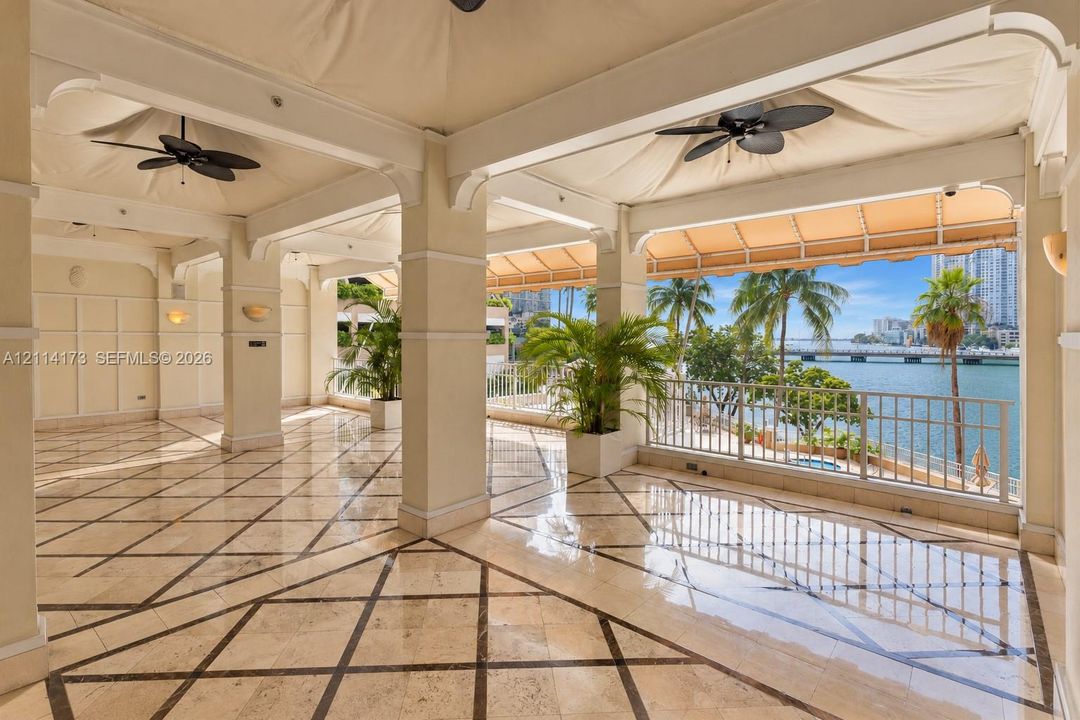 Covered terrace featuring stone tile flooring with decorative inlay, structural columns, ceiling-mounted fans, and metal railings overlooking the water