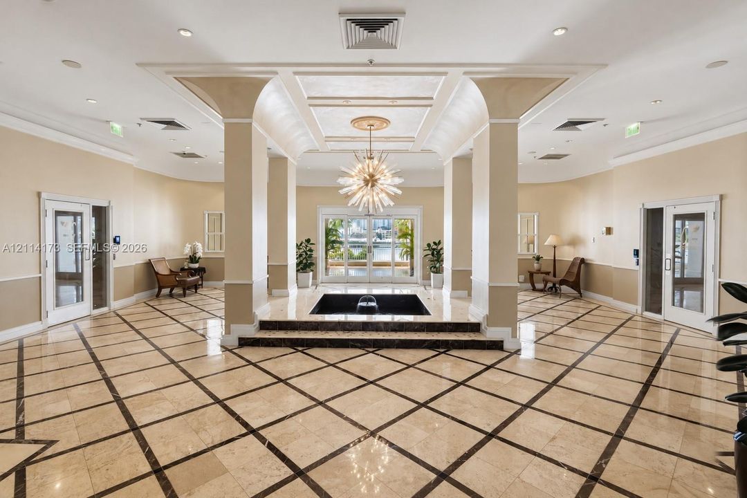 Grand lobby featuring polished stone flooring with geometric tile inlay, a central tiered water feature, soaring architectural pillars, recessed tray ceiling detailing, and an ornamental tiered light fixture