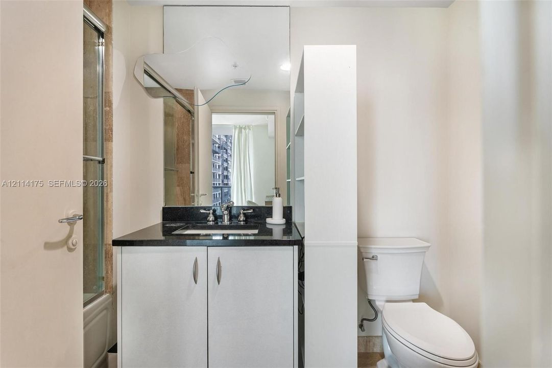 Vanity featuring white cabinetry, a dark stone countertop, integrated sink, wall-mounted mirror, and attached shelving unit