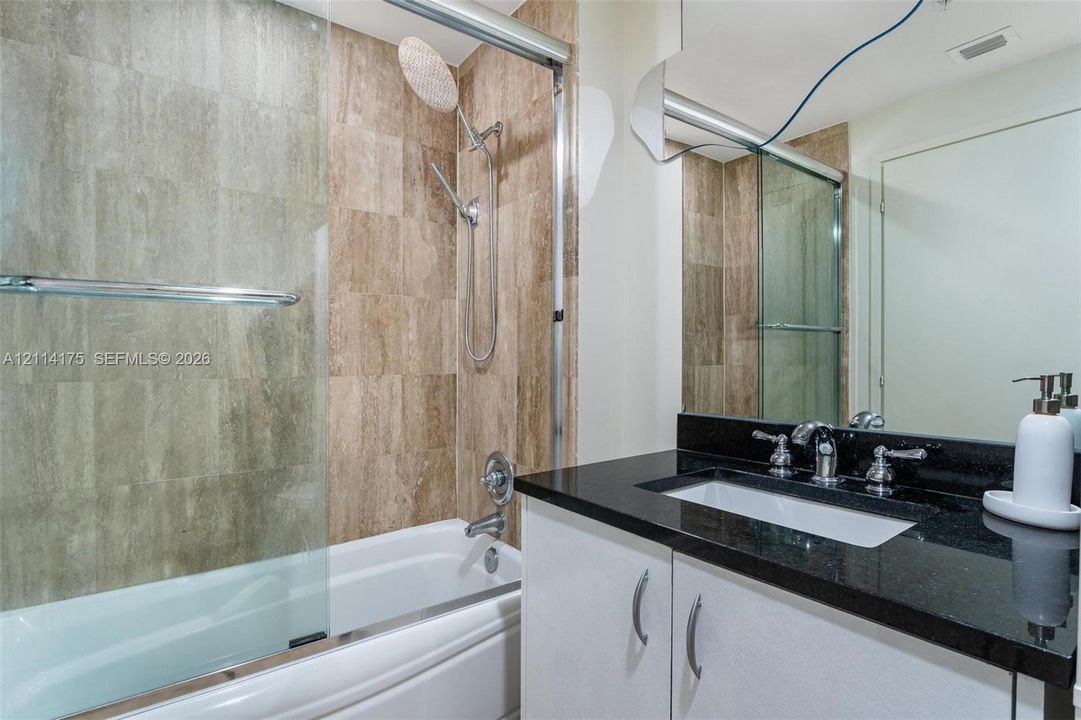 Bathroom featuring a vanity with dark stone countertop, an undermount sink with chrome dual-handle faucet, a shower-tub combination with full-height stone-finish wall tile, and a glass shower panel