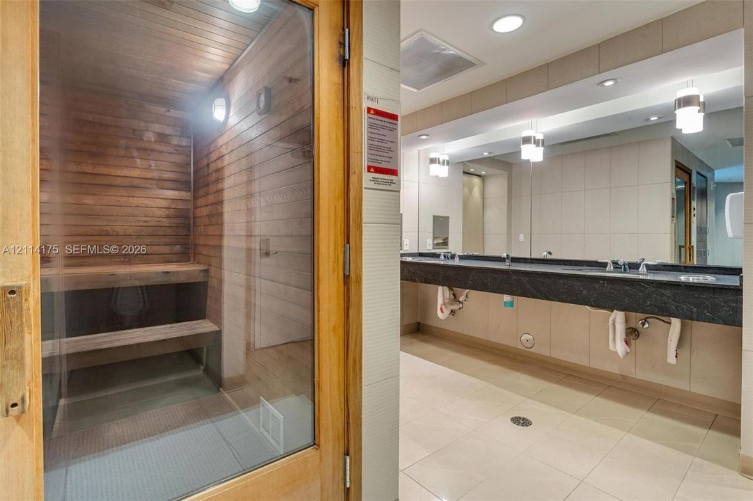 Wood-paneled sauna featuring a glass entry door, multi-level built-in bench seating, a wall-mounted vanity with a dual-basin countertop, and wall-to-wall mirrors