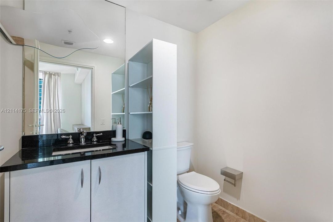 Bathroom featuring a vanity with dark stone countertop, integrated undermount sink, large wall-mounted mirror, built-in shelving unit, and white cabinetry