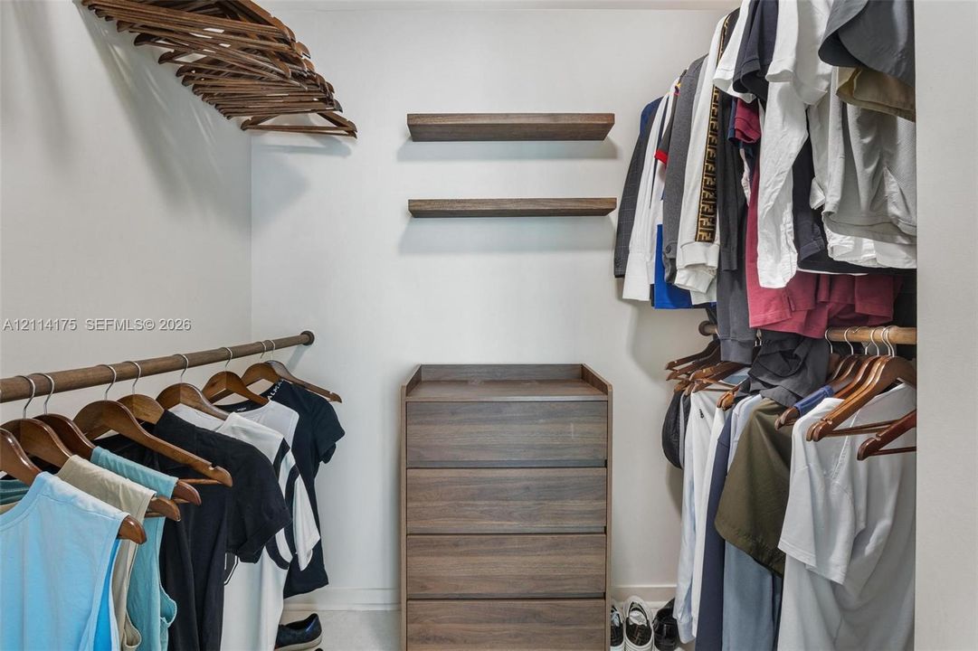 Walk-in closet featuring dual wood-finish hanging rods, two built-in floating shelves, and a four-drawer wood-finish dresser