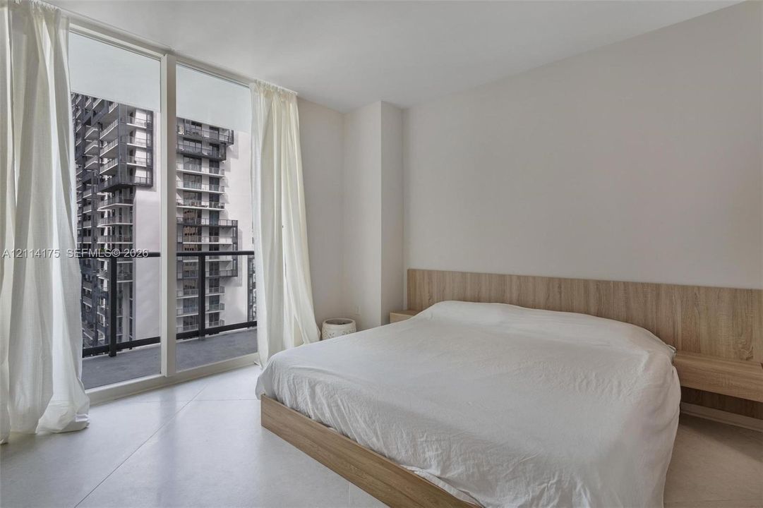 Floor-to-ceiling windows, integrated wood-finish headboard with built-in nightstands, and private balcony access