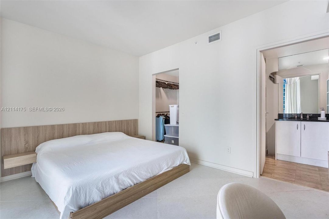 Built-in wood-finish headboard with integrated floating nightstands, stone-finish flooring, and vanity featuring a dark countertop and white cabinetry