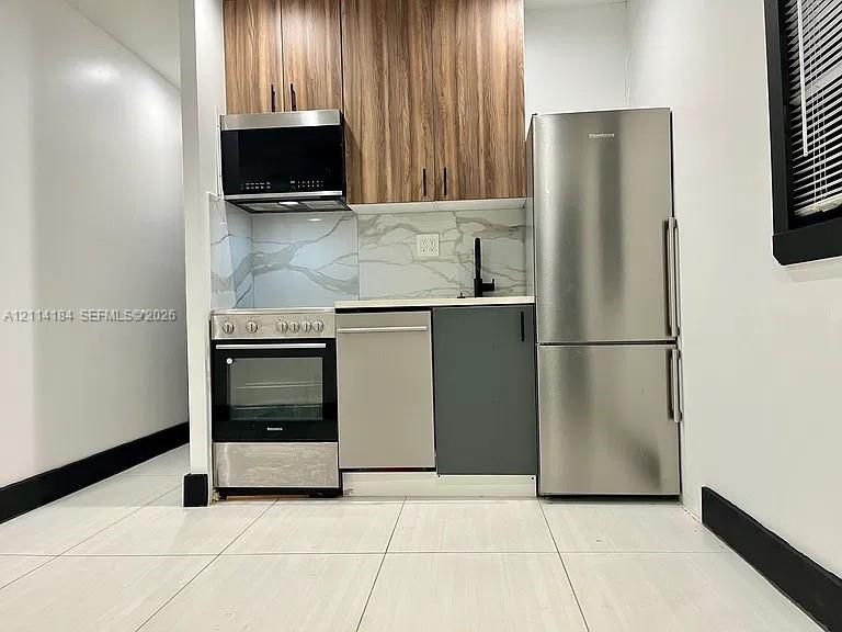 Kitchen featuring wood-finish upper cabinetry, stone-patterned backsplash, stainless steel appliances, matte black hardware, and large-format tile flooring
