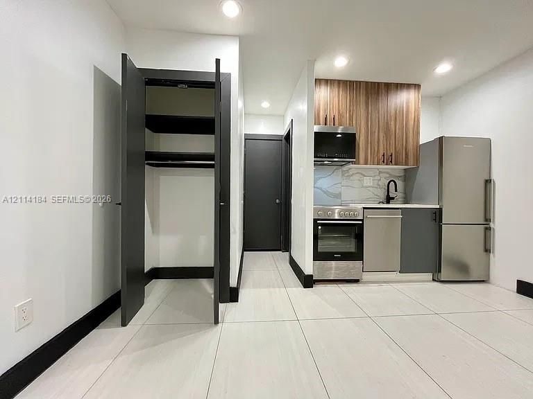 Kitchenette featuring dual-tone cabinetry, stainless steel appliances, stone-patterned backsplash, and large-format tile flooring, complete with a built-in storage closet
