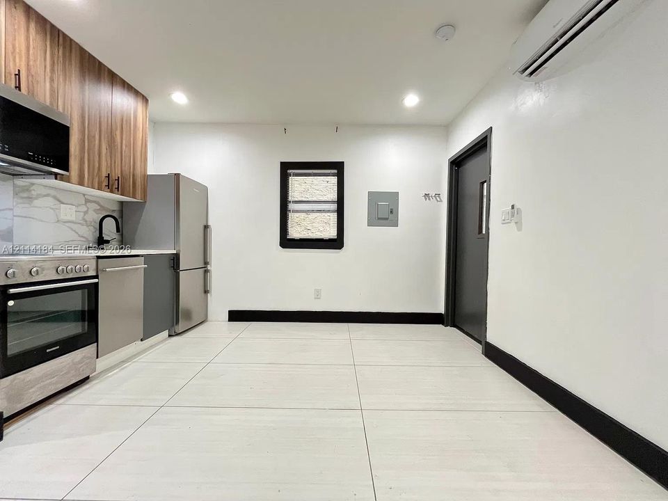 Kitchen featuring wood-finish cabinetry, stone-patterned backsplash, stainless steel appliances, large-format tile flooring, and a wall-mounted ductless heating and cooling unit