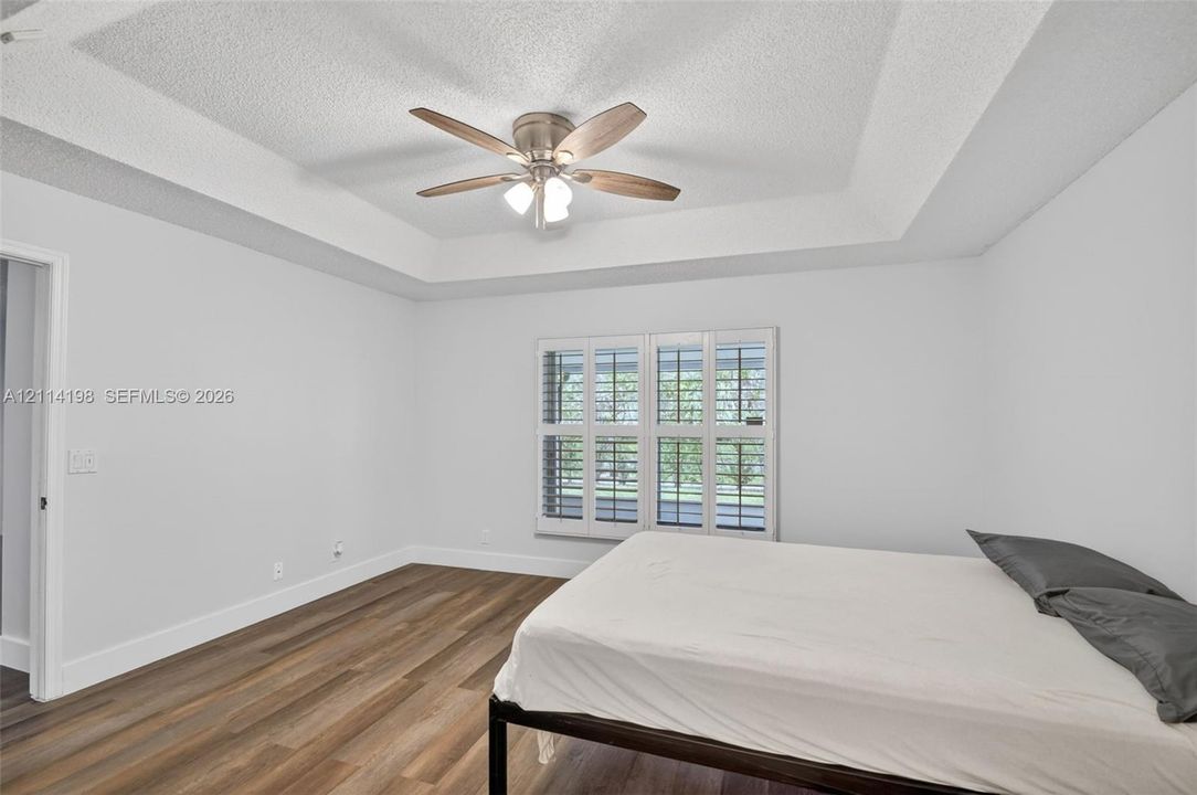 Room featuring a tray ceiling, wood-finish flooring, multi-pane window with interior shutters, and a central ceiling fan with integrated lighting
