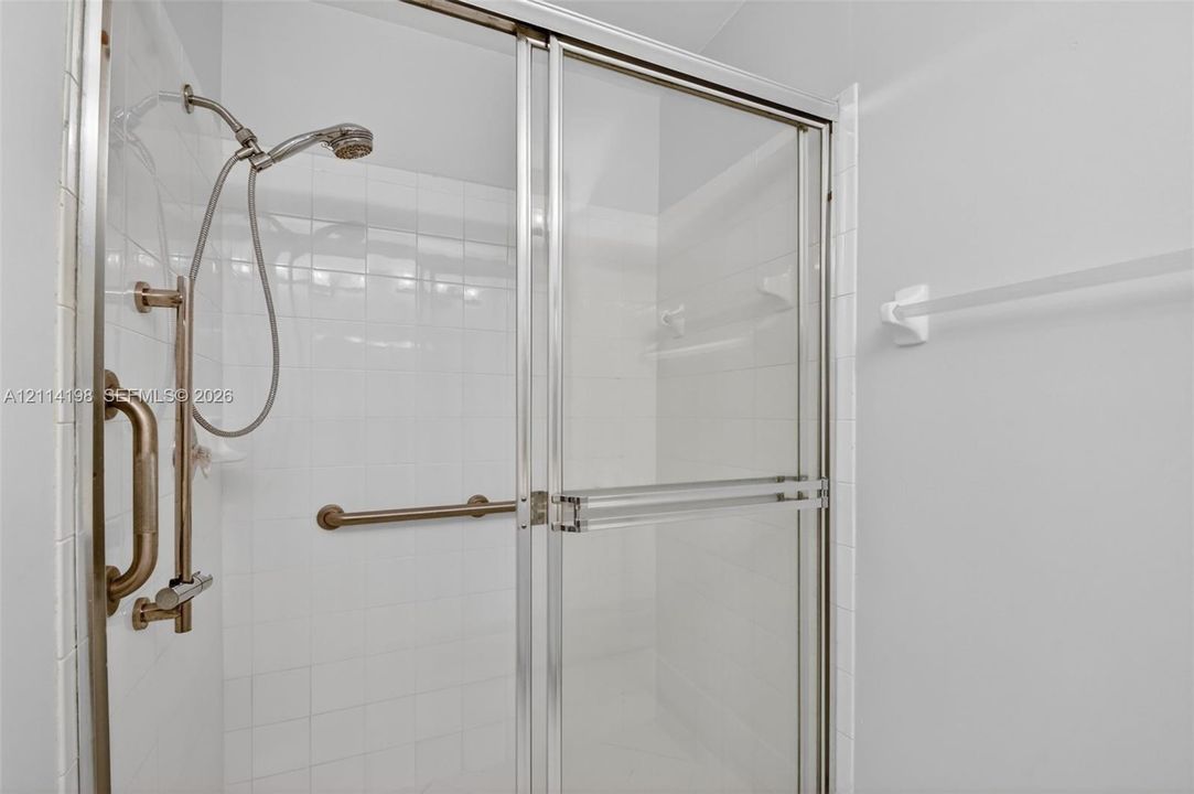 Walk-in shower enclosure featuring white tile wall surrounds, sliding glass doors, a handheld shower fixture on a slide bar, and metal safety grab bars