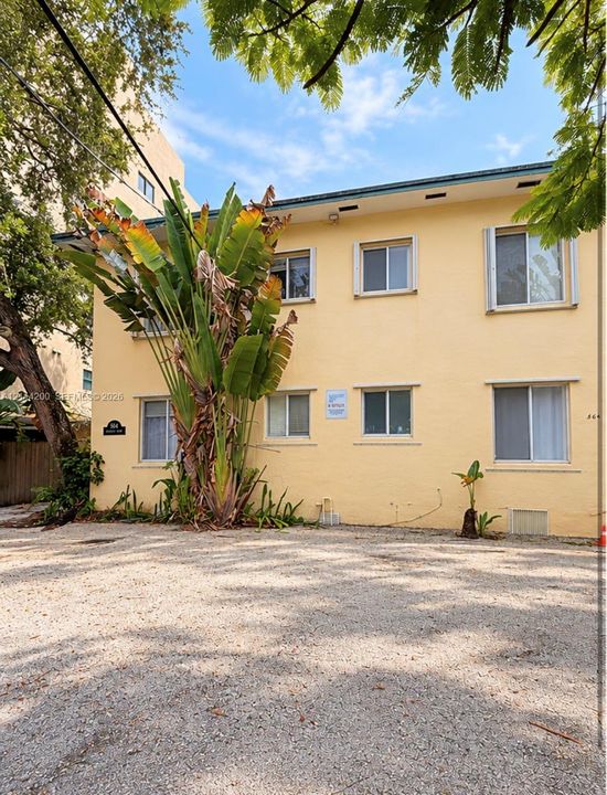Two-story multi-unit exterior with a light stucco finish, featuring sliding windows, mature palm landscaping, and an expansive gravel surface area