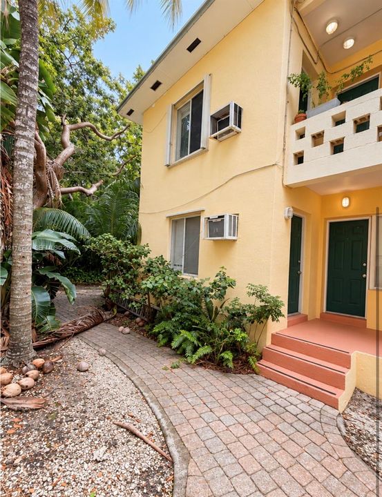 Two-story exterior featuring a stucco facade, second-level balcony with a concrete decorative screen, paved stone walkway, and external staircase