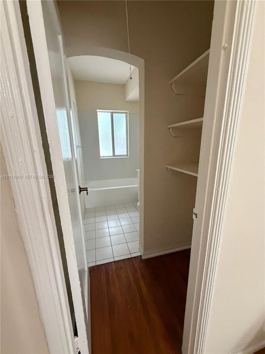 Storage area featuring built-in shelving, wood-finish flooring, and arched entryway leading to a bathroom with tile flooring, a window, and a bathtub