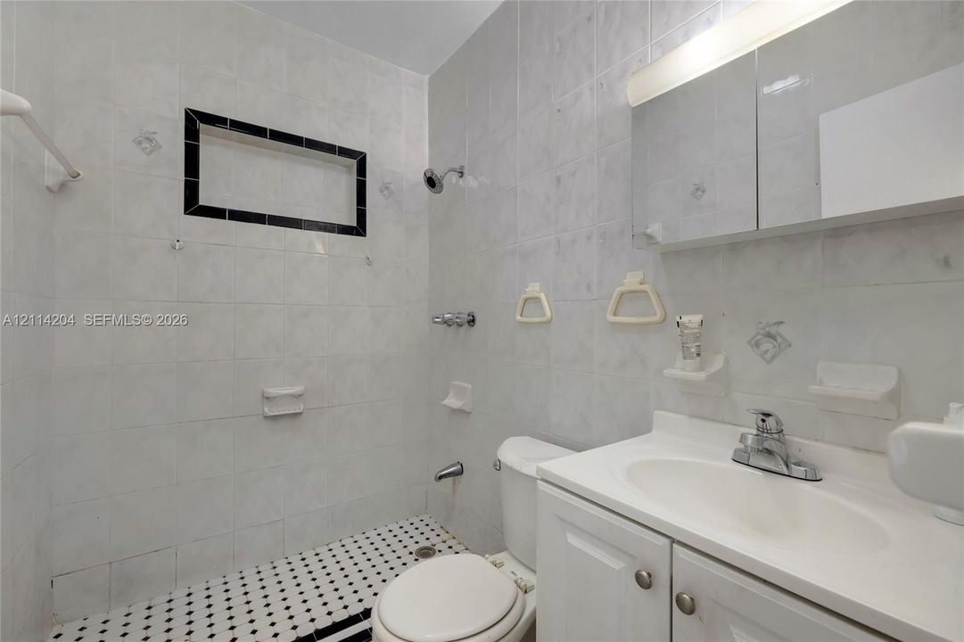 Full-bathroom featuring white wall tiling, an integrated wall niche with decorative black trim, a built-in medicine cabinet with vanity lighting, and black-and-white patterned floor tiling