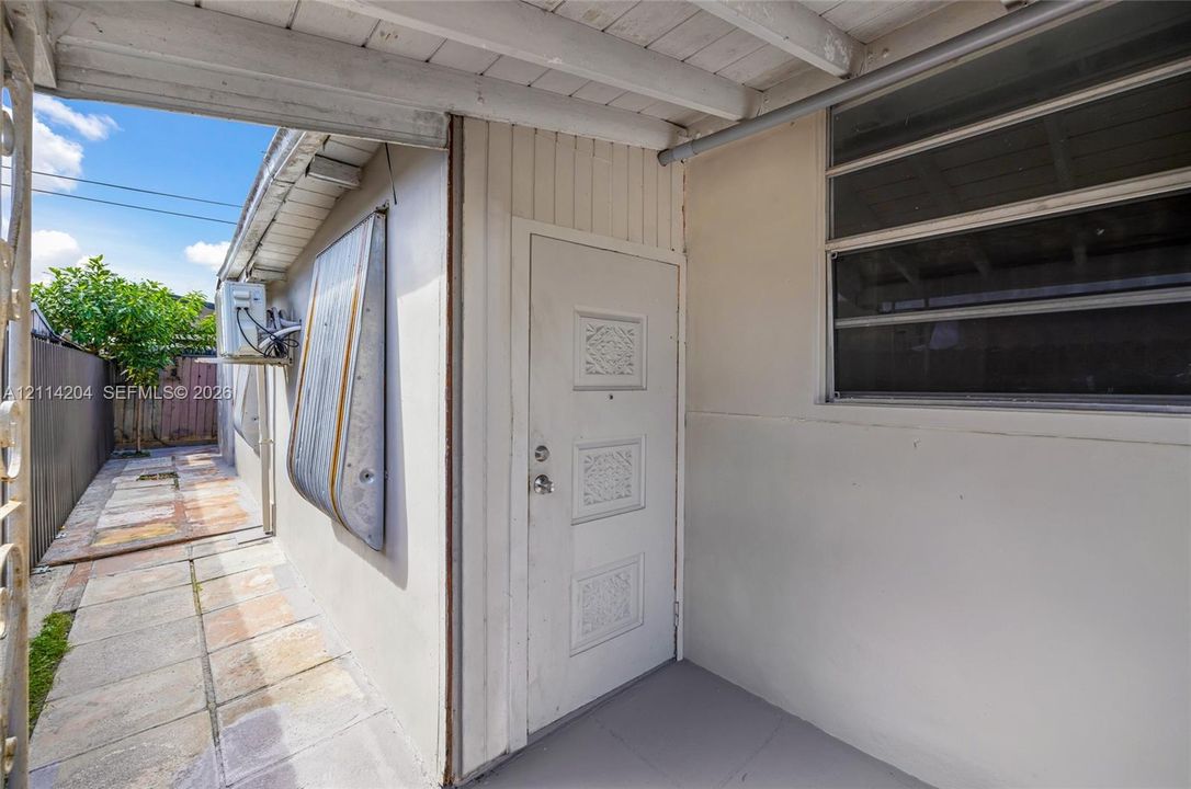 Covered exterior entry featuring a textured door, horizontal sliding windows, and paved ground surface