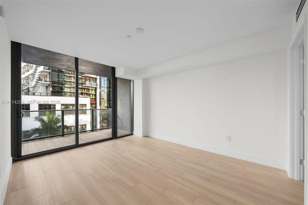 Floor-to-ceiling sliding glass doors, neutral wood-finish flooring, and an attached balcony with black metal railings