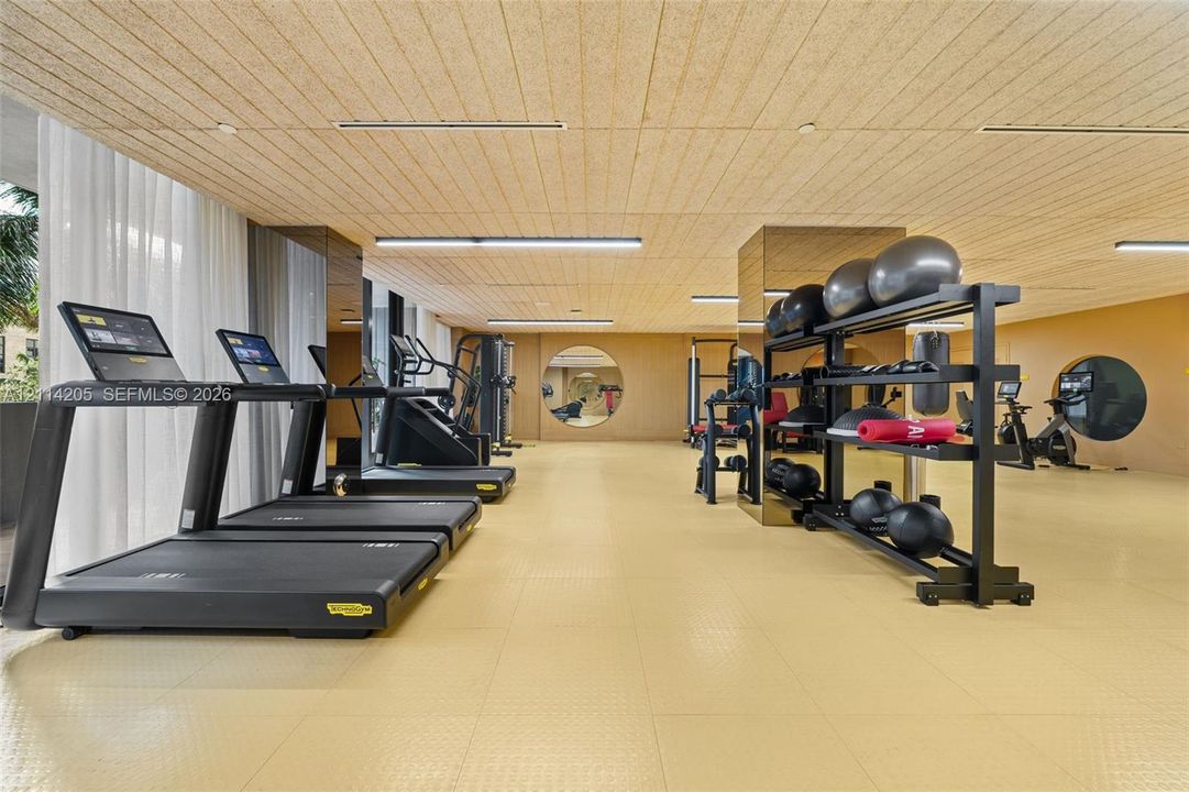 Fitness area featuring textured wood-panel ceiling, mirrored structural pillars, circular accent mirrors, recessed linear lighting, and resilient athletic flooring