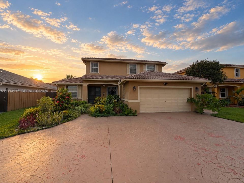 Two-story residence featuring a tile roof, stucco exterior, attached two-car garage, and a decorative stamped concrete driveway surrounded by established landscaping