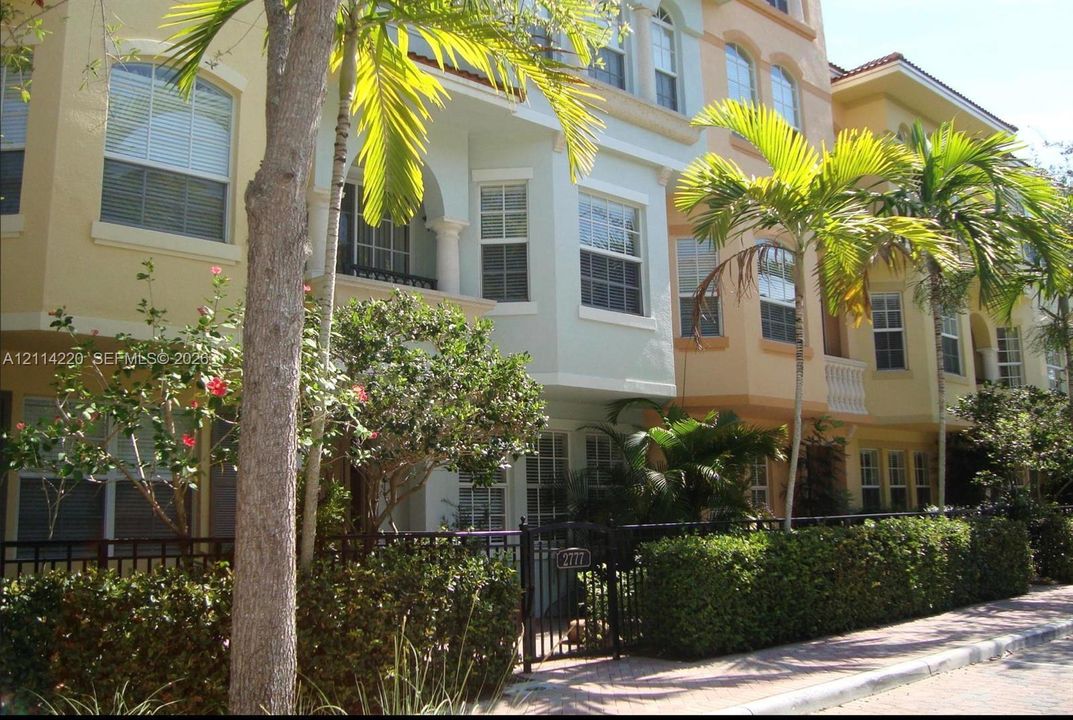Multi-story exterior featuring a light-toned stucco façade, arched window detailing, wrought iron fencing, and established palm landscaping