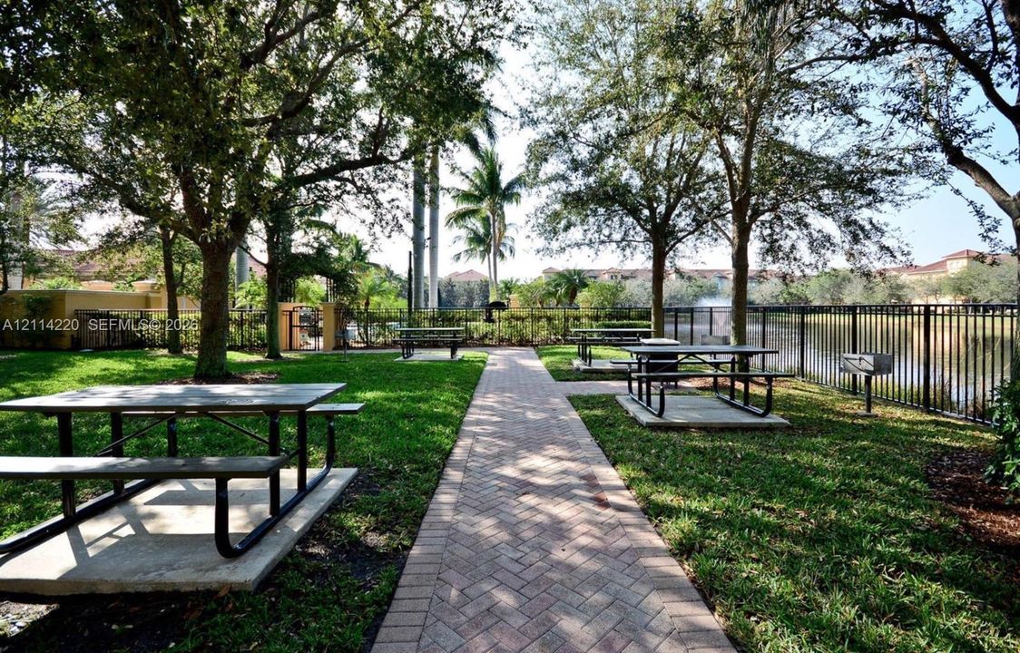 Outdoor recreation area featuring a brick-paver walkway, multiple built-in metal picnic tables on concrete pads, manicured lawn, mature trees, and a perimeter metal fence bordering a water feature
