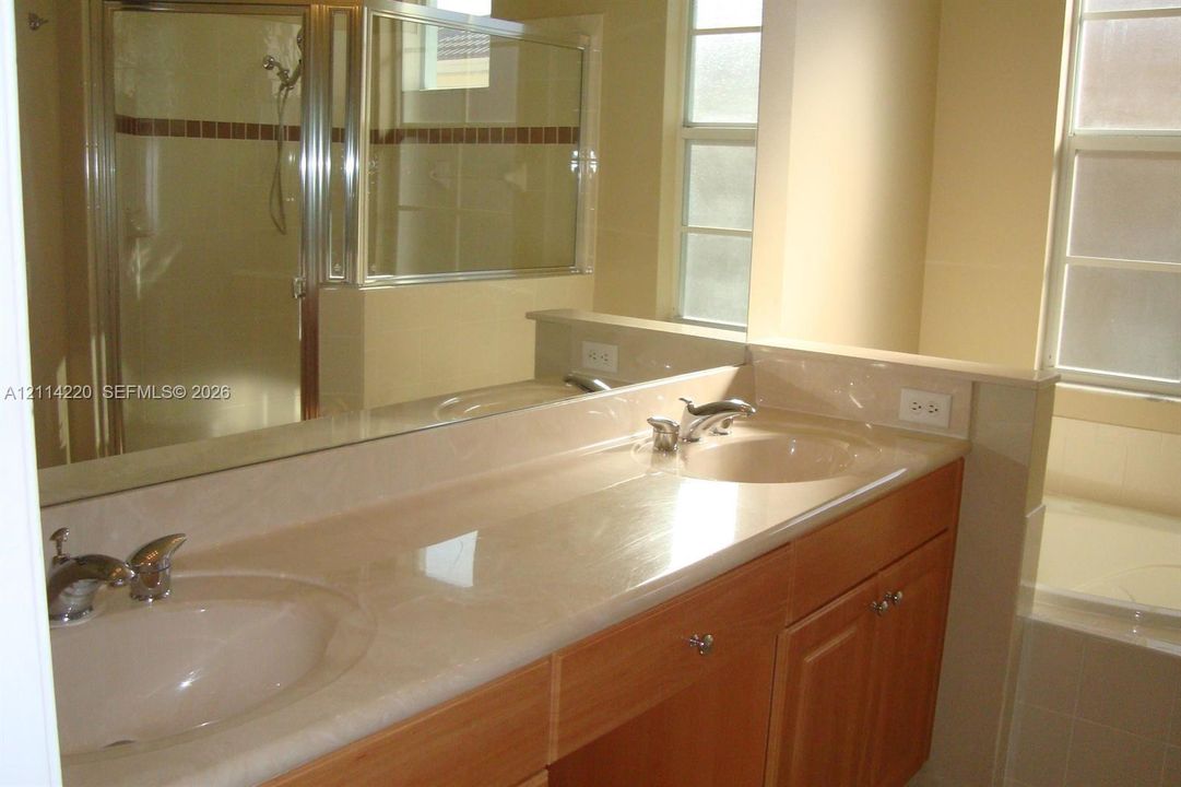 Dual-sink vanity with wood-finish cabinetry, solid surface countertop, chrome hardware, separate glass-enclosed shower, and built-in soaking tub