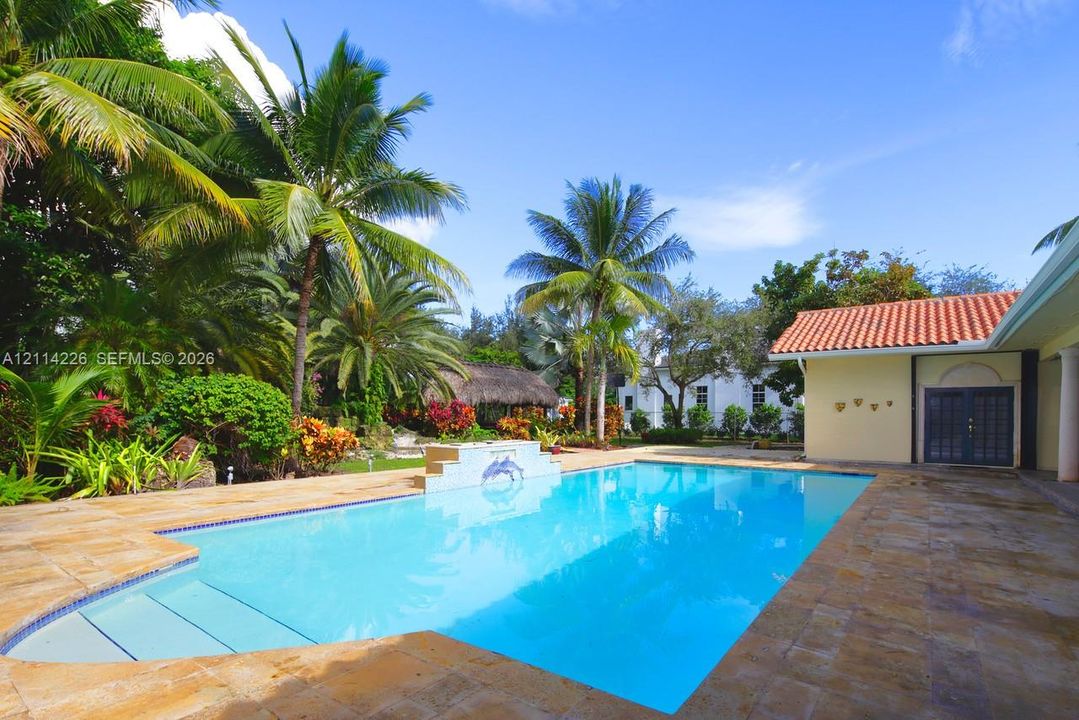 In-ground swimming pool with built-in water feature and tiled steps, surrounded by stone-paver decking and mature tropical landscaping