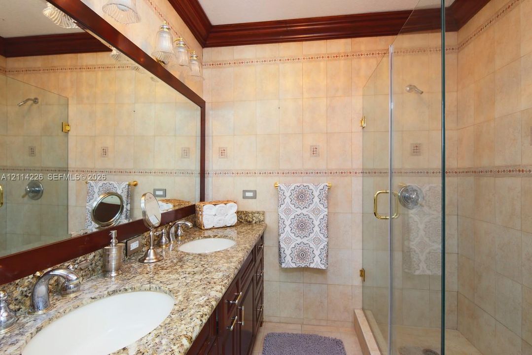 Bathroom featuring a granite countertop vanity with dual undermount sinks, custom dark wood cabinetry, a glass-enclosed shower with tiled walls, and crown molding