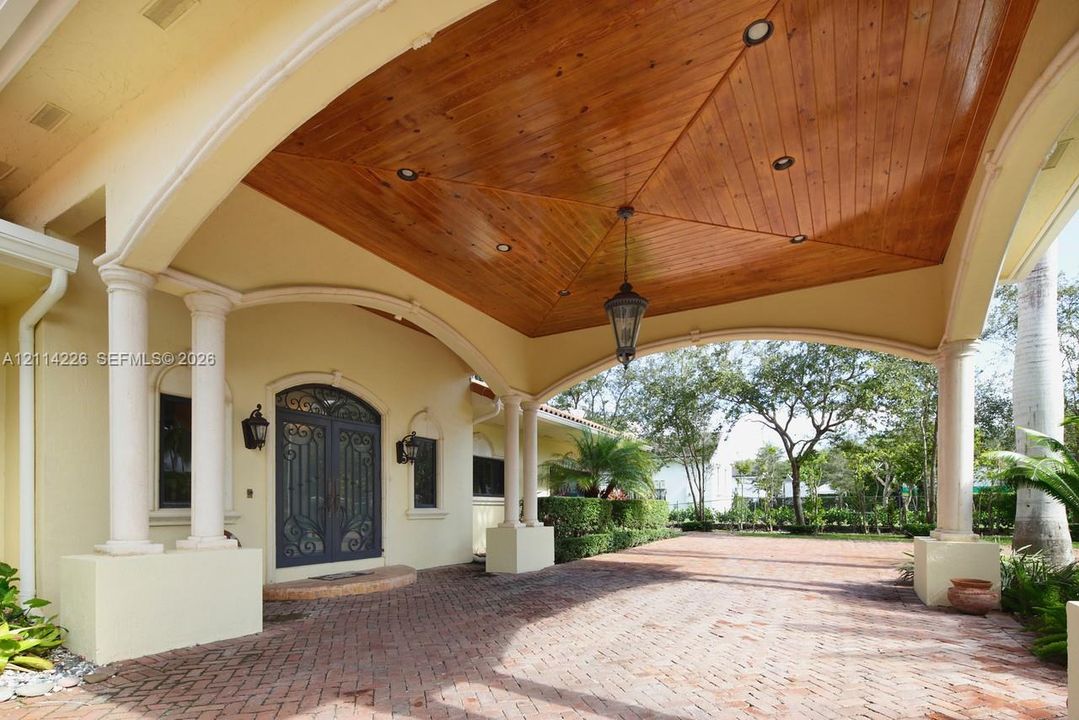 Covered porte-cochère featuring a wood-plank vaulted ceiling, recessed lighting, ornate double front entry doors, and brick paver flooring supported by classical columns
