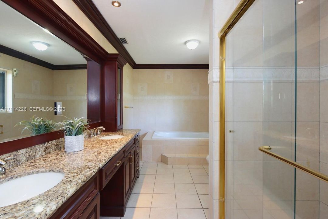 Bathroom featuring a double-sink vanity with dark wood cabinetry and stone-surface countertops, a deep soaking tub with tile surround, a walk-in glass-enclosed shower with gold-tone hardware, and floor tiling throughout