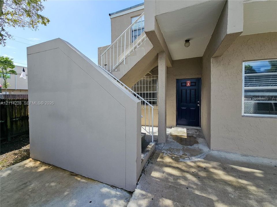 Exterior entry featuring stucco siding, solid-panel door, concrete porch, and metal stair railing