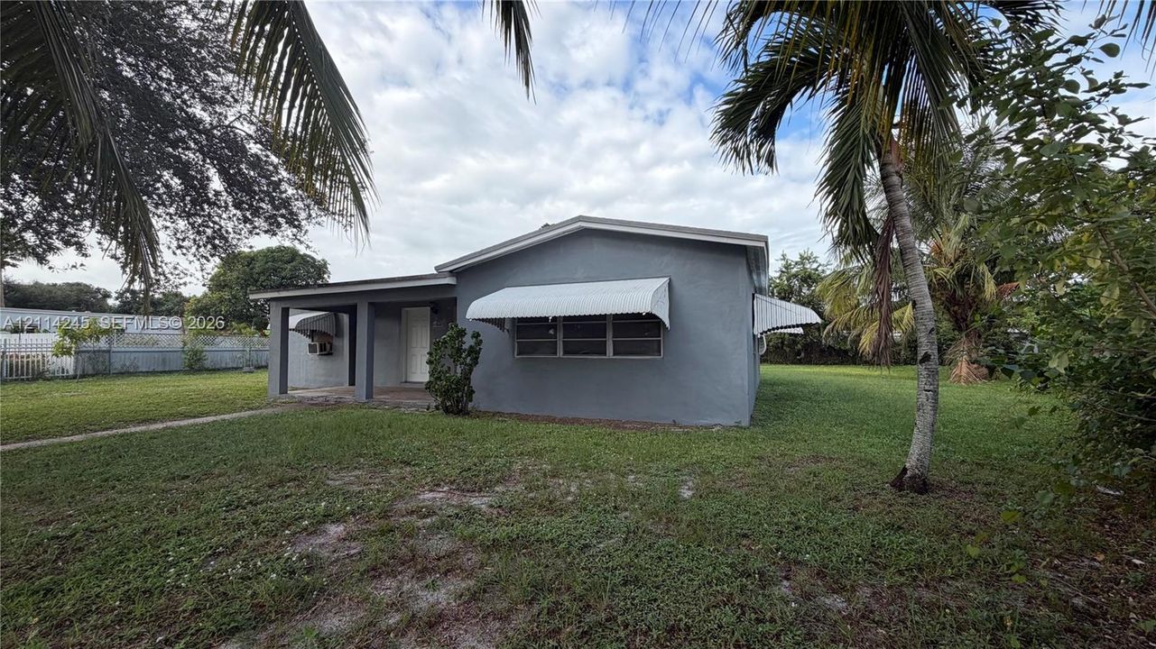Exterior facade featuring a stucco finish, covered entryway, metal window awnings, and expansive lawn area with mature palm trees