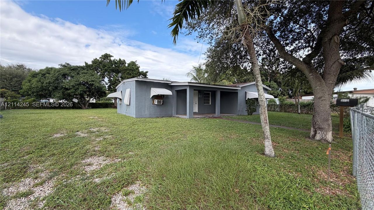 Single-story residence featuring a stucco exterior, covered front porch, metal window awnings, and a chain-link perimeter fence on a spacious lot with mature trees