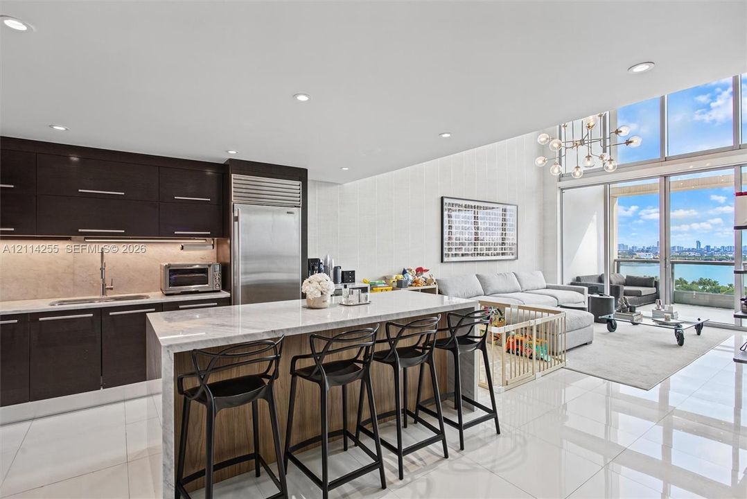Kitchen featuring dark custom cabinetry, stone-topped waterfall island, professional-grade stainless steel refrigerator, recessed lighting, and floor-to-ceiling windows