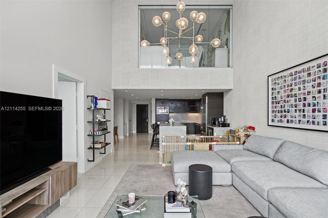 Double-height ceiling, large multi-globe chandelier, polished tile flooring, kitchen island with stone-finish waterfall edge, and textured wall finish