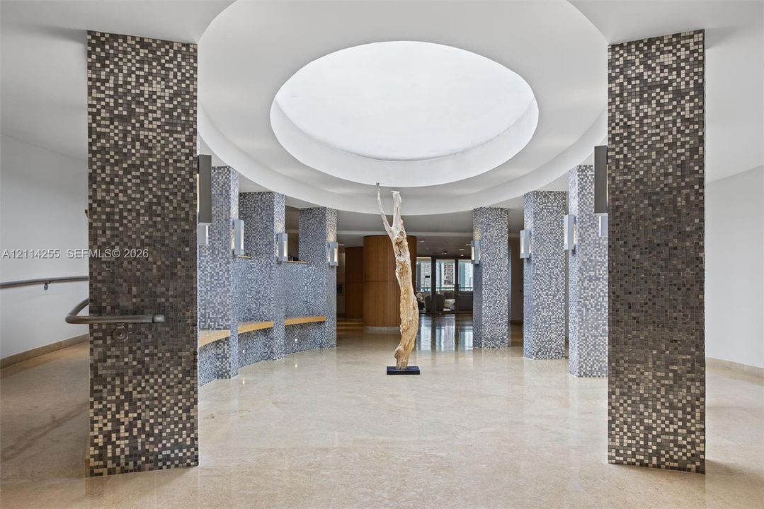Circular rotunda featuring a recessed dome ceiling, mosaic-tiled structural columns, built-in wooden benches, and polished stone flooring