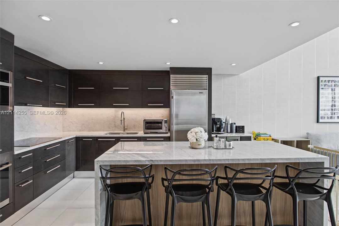 Kitchen featuring dark wood-finish cabinetry, a waterfall-edge stone island, under-cabinet lighting, stainless steel appliances, and large-format tile flooring