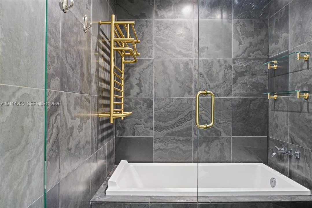 Large-format charcoal stone tile surround with a white deep-soaking tub, brass-finish heated towel rack, wall-mounted faucet, and glass shelving