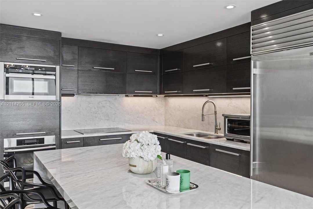 Custom dark-finish cabinetry featuring horizontal bar pulls, expansive stone-surface center island, integrated stainless steel refrigeration, built-in wall oven and microwave combination, and stone-surface backsplash with under-cabinet lighting