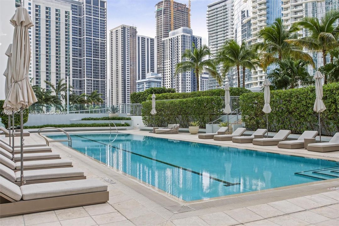 Swimming pool with a lap lane, surrounded by stone-tile decking and mature privacy hedging, featuring a prominent urban skyline backdrop