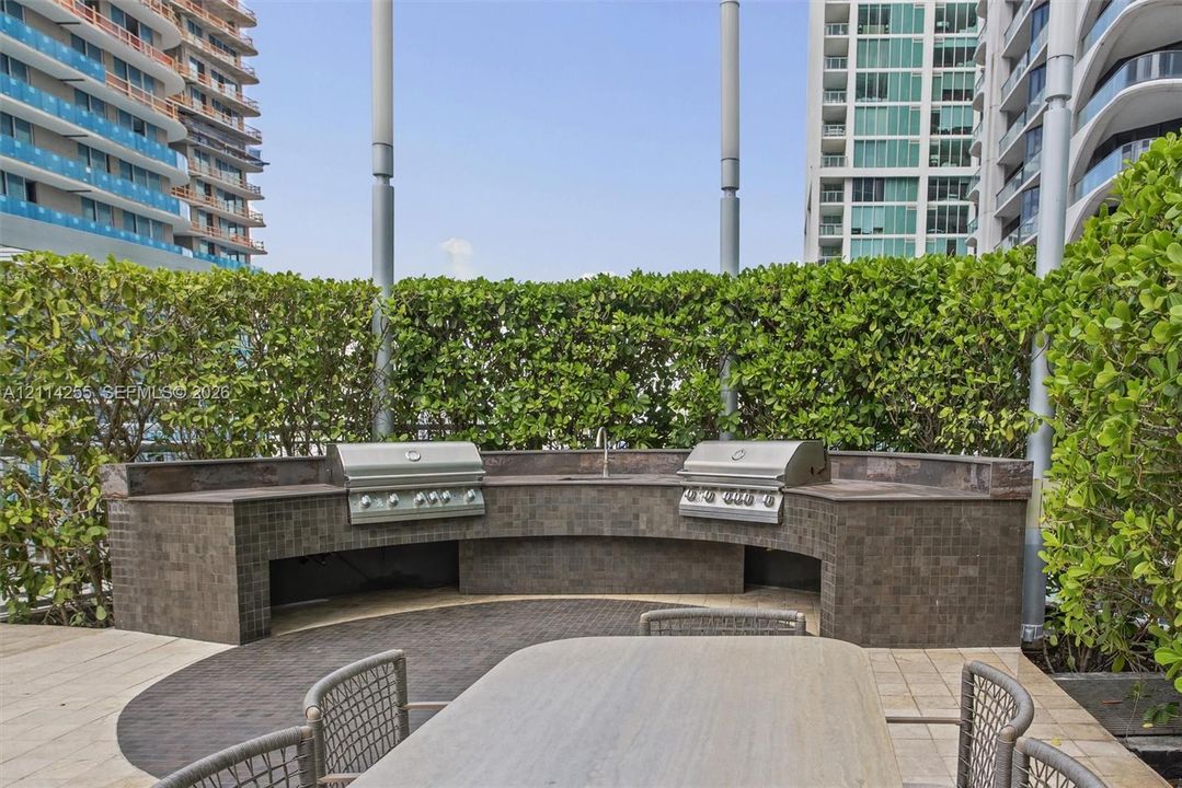 Curved outdoor kitchen featuring dual stainless steel grills, a stone-tiled backsplash, and built-in prep counters, framed by integrated perimeter hedges